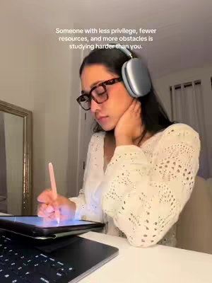 A well-lit, indoor shot featuring a woman with dark hair and glasses. She is holding a pen, appearing to be writing or drawing on a tablet resting on a desk, with a laptop keyboard visible below. The style is minimalistic and focus is on productivity and studiousness.