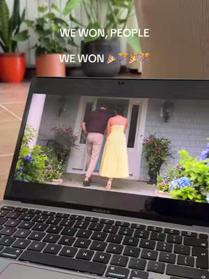A laptop displays a scene from "The Summer I Turned Pretty" with a couple walking towards a house. Above the screen, the text "WE WON, PEOPLE\nWE WON" in white font, along with butterfly and sparkle emojis, floats against a background with plants. The laptop is open on a wooden surface, with a focus on the screen featuring the show's scene. The background includes visible indoor plants.
