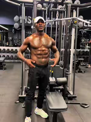 A muscular man stands shirtless in a gym, flexing his abs. He wears black pants, white shoes, and a white cap. Gym equipment is visible in the background.