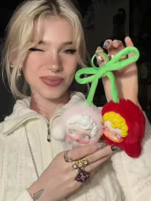 A woman with blonde hair is holding two plush toys shaped like cherries. The woman is smiling and winking. The plushies are white and red with green stems.
