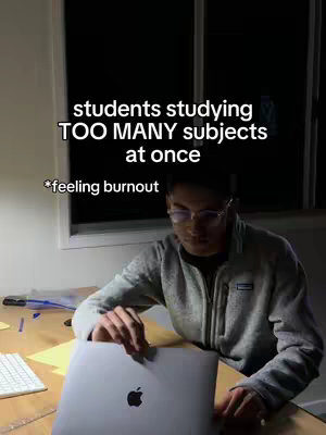 A dimly lit interior shot featuring a student seated at a desk, working on a laptop. The background is dark, with a window letting in minimal light. The student wears eyeglasses and a casual gray hoodie. Overlay text at the top reads, "students studying TOO MANY subjects at once." Another line of text at the bottom says "feeling burnout."