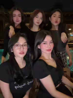 Five young women pose closely together, smiling at the camera. The setting appears to be outdoors, with a pool visible in the background. The women are dressed in dark clothing, and the lighting suggests it might be dusk or evening. The overall composition focuses on their faces and upper bodies, creating a sense of camaraderie.