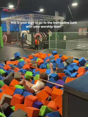 A person wearing a white shirt is submerged in a foam pit filled with colorful blocks. The image is taken indoors. In the background is a metal structure and other people. Text overlay indicates the location is a trampoline park.