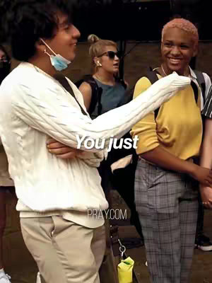 A group of people are standing on a street talking. The main focus is on a young man on the left, who seems to be gesturing towards the other two. The man on the left has a white sweater and a mask under his chin, talking to a woman on the right wearing a yellow shirt giving a welcoming smile.
