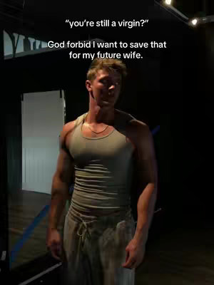 A man in a gray tank top and loose pants stands in a dim room, looking slightly to the side. Text overlays appear above him.