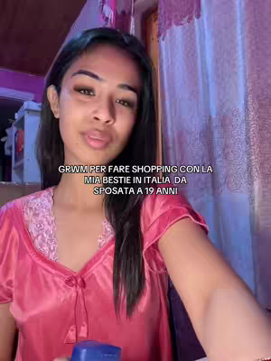 A young woman with long dark hair and a soft expression poses for a selfie. She's wearing a pink satin top and a hint of makeup. The background suggests a bedroom setting with curtains.