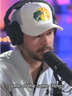 A man with a mustache and a white cap is holding a microphone. The man has headphones on and is positioned in front of a blurred purple and blue background.