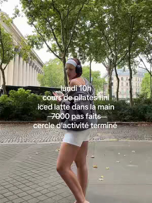 A woman is walking on a city street, holding a drink and wearing headphones. Text is overlaid on the image detailing her activities, including a pilates class, steps taken, and an activity circle.