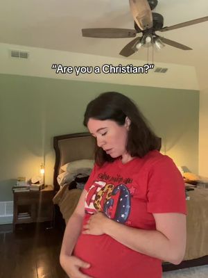 The image shows a woman in a domestic setting, likely a bedroom. She is pregnant, indicated by her prominent belly, and is wearing a red t-shirt with a graphic. She appears to be looking down and cupping her belly with her hands, which conveys a sense of tenderness. The background includes a bed with rumpled bedding, a nightstand, and a ceiling fan. The lighting is soft. A text overlay in a speech bubble reads: Are you a Christian?