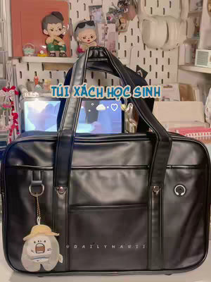 A black leather bag in the foreground, possibly a school bag. The bag has a front pocket and is hanging with a gray cartoon character charm. Behind the bag is a cluttered shelf with cute figurines and stationery, creating a cozy aesthetic.