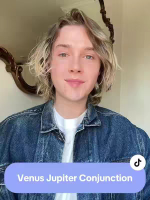 A selfie-style image featuring a young man with light brown hair. He's wearing a denim jacket over a white t-shirt. In the background, there's a glimpse of a mirror and what appears to be a wall. A light blue oval-shaped text box at the bottom of the image reads "Venus Jupiter Conjunction". The overall composition is centered and framed from the chest up.