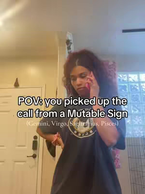 The cover image is a medium shot of a person on the phone, appearing to be indoors, possibly within a home setting. The person, who seems to be female, is wearing a dark t-shirt and has long hair. They are holding a pink phone to their ear and their expression suggests mild exasperation. Text overlay indicates the video's focus on astrology and mutable signs. The setting includes a white door, a portion of a glass block window, and other room elements.