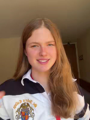 A young woman with long, light brown hair smiles at the camera. She is wearing a white rugby shirt with navy and black details and the emblem of a college. The background is a light-colored wall with a door and a window.