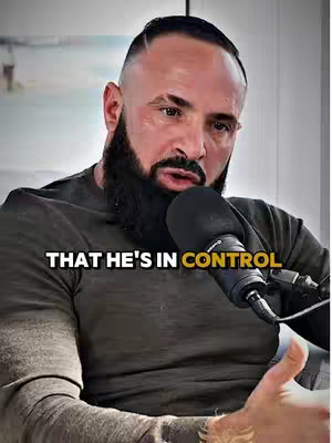 A close-up shot of a man with a shaved head and a full beard, wearing a dark olive-green t-shirt, speaking into a black microphone. The text overlay reads \"THAT HE'S IN CONTROL\". The background is slightly blurred out, suggesting an indoor setting possibly a living room or a studio set.