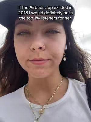 A selfie-style video with a young woman in the frame. The woman is fair-skinned, with dark, wavy hair. She is wearing a dark hat, a white shirt along with gold accessories and a necklace. The background is blurred with an overcast sky. A text overlay is present above her face, with the text in a white font. The overall composition is focused on her face and upper body.