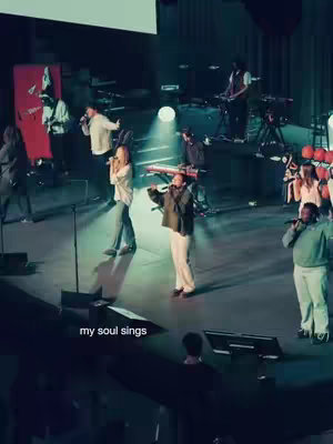 A live performance on stage, featuring several people singing and playing instruments. A group of people are on stage, with the central focus on three people holding microphones, singing. The lighting is dim with a prominent spotlight shining on the performers. The background includes stage equipment and a large screen.