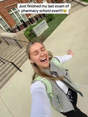 A selfie taken from a slightly low angle, featuring a young woman with blonde hair and a bright, joyful expression. She is standing outside a brick building with large windows, and a sign is visible in the background. The woman is wearing a light green vest over a white shirt and carrying a backpack. Her arms are outstretched, and her mouth is wide open in a laugh, suggesting excitement or relief. The overall composition is well-lit, emphasizing the celebratory mood.