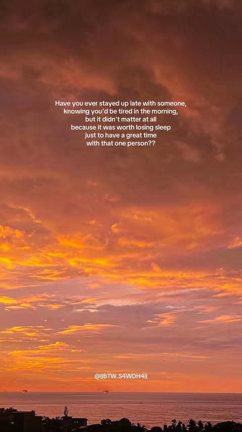 A vibrant sunset over a body of water, possibly the ocean. The sky is filled with rich, warm colors, transitioning from orange to yellow, with hints of purple and pink in the clouds. The water reflects the colors of the sky. The foreground has a dark silhouette, likely of buildings or land. Text is overlaid in the sky.