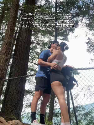 A couple is kissing in a natural setting. The image is taken from a slightly low angle, emphasizing the trees in the background that frame the couple kissing. The composition is candid, with the subjects in focus and surrounded by blurry trees.