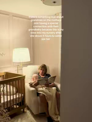 The image shows an interior scene with a grandmother and a baby. The grandmother, a woman with short blonde hair, is seated in a white armchair, holding the baby in her arms. They are both looking at a book. Behind them, a baby crib made of wood is visible. The room has white walls and a beige carpet, and the lighting suggests a warm and cozy setting.