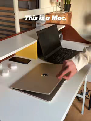The image shows a person opening a laptop, with another laptop visible on the desk, implying a comparison. The setting is a room with a bed or shelf in the background.