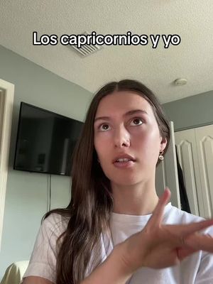 A woman with long, dark hair, dressed in a white t-shirt, is looking upwards with a thoughtful expression. Her right arm is raised with fingers gesturing outward. Above her is text that reads ' Los capricornios y yo'. The background shows an interior with a wall, a closed door, and a large dark-screened television.