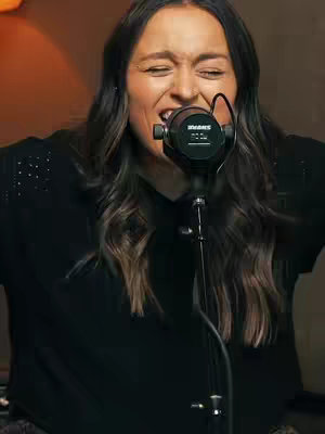 A woman with long dark hair, wearing a black top sings into a microphone with her eyes closed. The background is softly lit in warm tones.