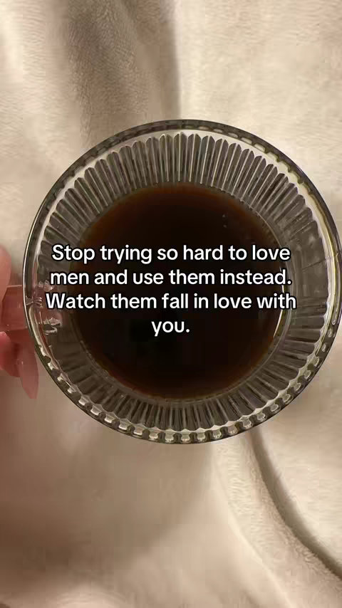 A close-up shot of a glass of coffee. In the foreground, a clear glass cup filled with dark coffee is visible. The cup is held on a white surface, presumably a bedsheet or blanket, with a hand visible holding the cup. Written overlay text is in the center of the cup and reads, “Stop trying so hard to love men and use them instead. Watch them fall in love with you.” The background is a soft, blurred white, which appears to be a bed.