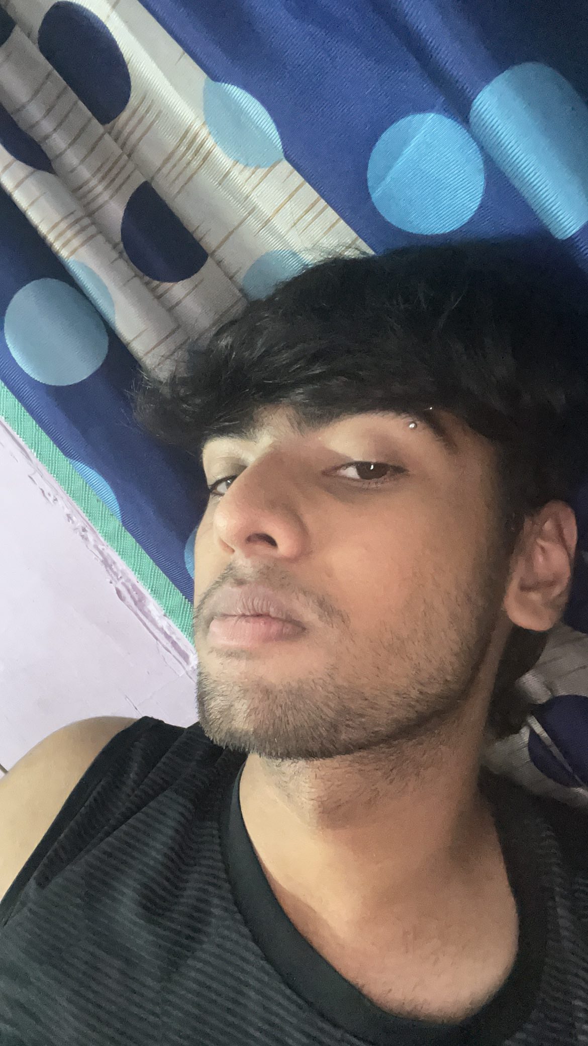 A selfie-style shot shows a young man with dark hair, wearing a black tank top. He has a small eyebrow piercing and a closely-shaved beard. The background consists of a blue and white patterned curtain, and the lighting appears to be natural.