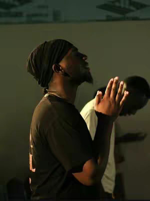 A medium shot focuses on a Black man wearing a black beanie and a black t-shirt, with his hands clasped in prayer and eyes looking upward. Another person in a white shirt is slightly blurred in the background, also with their hands in prayer. The lighting suggests an indoor setting.