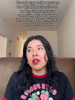 A woman with long black hair, red lipstick, and a black t-shirt with a strawberry design looks up with an expressive face, seemingly talking. Text overlayed on the image discusses a partner with Venus in Leo, suggesting a relationship theme. Behind her are neutral-toned walls and a glimpse of a brown couch.