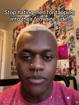 A close-up shot of a person with short, light-colored hair and a nose piercing, wearing a red shirt. The background is a wall covered in framed images and photos. The text 'Stop hating men for tapping into their feminine side' is overlaid at the top.