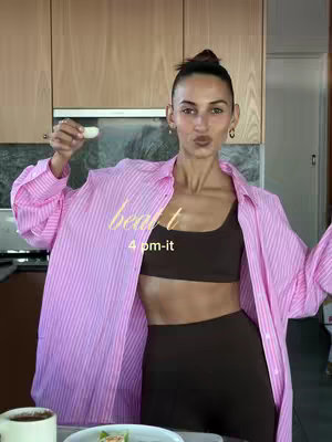 A woman in a pink striped shirt and brown athletic wear holds a small white object in a kitchen setting. Text overlays read 'beat' and '4 pm-it'. The background includes wooden cabinets and a partial view of the kitchen.