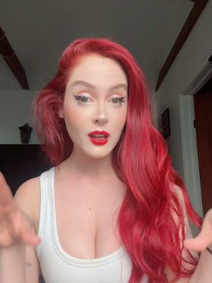 A woman with long, vibrant red hair and heavy makeup looks directly at the camera. She is wearing a white tank top, and her red lipstick is prominent. The background is a softly lit interior.