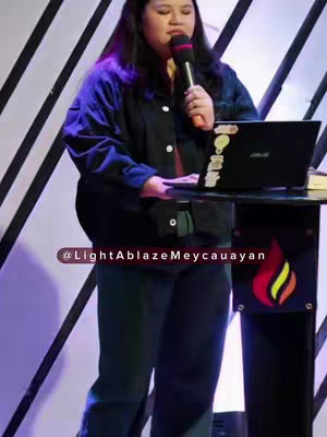 A medium shot shows a woman in a church setting, speaking into a microphone. She is positioned behind a podium with a laptop and a design of a flame. The background has vertical lines.