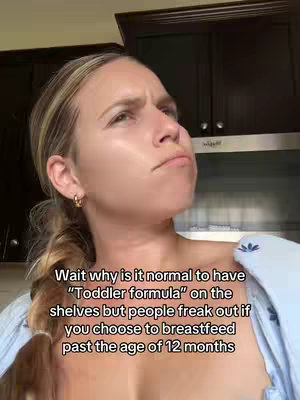 A selfie-style video cover image features a woman looking upwards with a questioning expression, her eyebrows furrowed. Her hair is blonde, pulled back in a ponytail, and she wears hoop earrings. Text on the image poses a question about the normalization of toddler formula versus the reaction to extended breastfeeding.