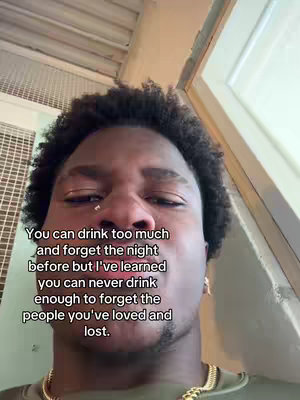 A close-up shot of a young Black man looking up at the camera, with text overlaid. The man has short, dark, curly hair and a small nose ring. He is wearing a gold chain and a dark green shirt. The background is slightly out of focus, including a metal grate and a section of a white wall and window trim. The text is centered and uses a white font.