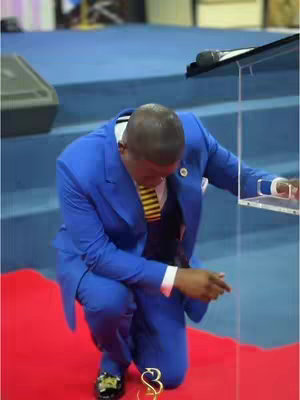A man in a blue suit kneels on a red carpet, leaning toward a transparent podium. A microphone is attached to the podium. in the background, there are some blue steps. His head is bowed.
