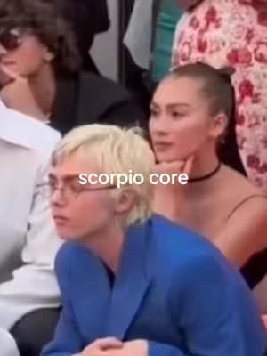 A candid, medium shot captures three individuals. A person with short blonde hair and glasses sits in the foreground, angled slightly toward the left. They wear a blue blazer. Beside them, a woman with dark hair pulled up in a bun and a black choker looks on, resting her face on her hand. Behind them, there are more people, blurred. The overlay text "scorpio core" appears in a white, sans-serif font.