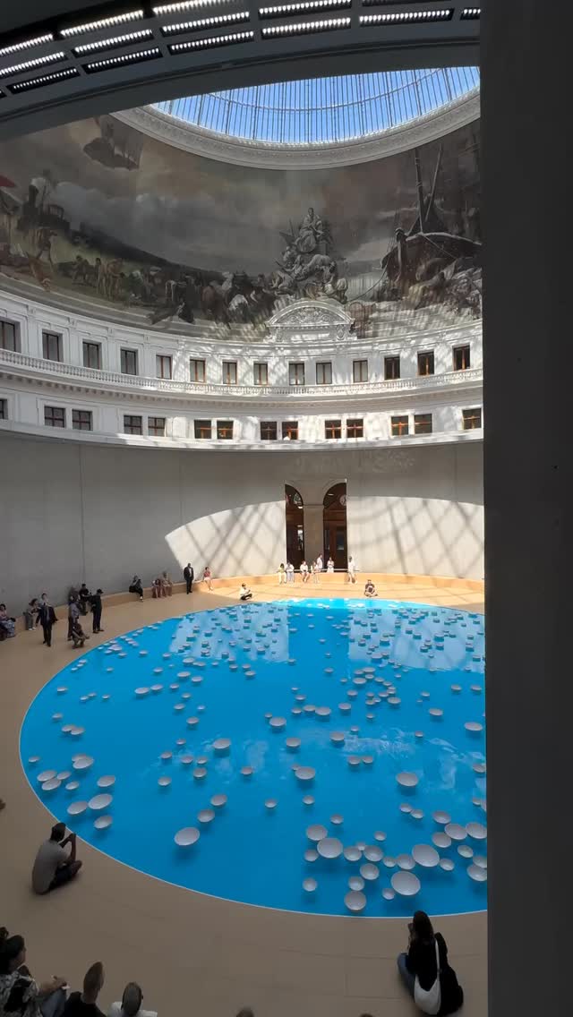 An interior shot of a large, circular building with a high, domed ceiling. The space features a large, turquoise-colored circular pool in the center, dotted with numerous white, disc-shaped objects. People are seen in the surrounding area. The ceiling has a mural.
