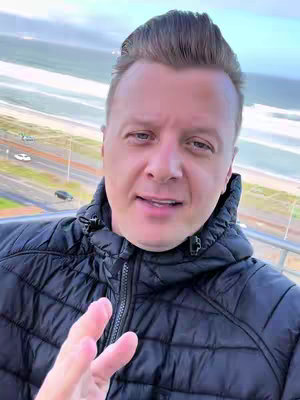 A man in a black jacket stands on a balcony, with a coastal scene in the background. He is looking towards the camera with an open expression, and his hand is up in a peace sign. The background shows a beach, ocean, and buildings along the coast under an overcast sky.