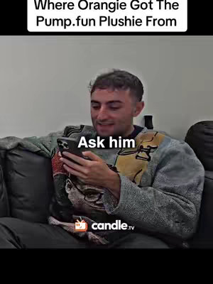 A man with short hair is sitting on a couch, holding a black smartphone and looking at it. The background is a light colored wall. Overlay text at the top says 'Where Orangie Got The Pump.fun Plushie From' and at the center of the image is the text 'Ask him'. The man is wearing a grey sweater with a detailed pattern.
