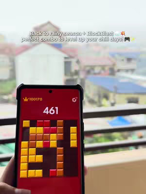 The cover image shows a smartphone displaying a Block Blast game, held by a hand. The background features a blurred view of buildings and foliage. Text overlay reads: “Back to rainy season + BlockBlast perfect combo to level up your chill days! 🎮.”