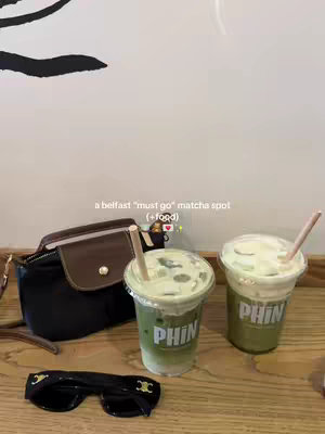The image showcases two matcha lattes in clear plastic cups with green liquid and white foam, placed on a wooden surface. Each cup has a paper straw and the brand 'PHIN' printed on it. Beside them are a black purse, sunglasses, and two black lines on a white-painted background. The text overlay reads 'a belfast "must go" matcha spot (+food)'.