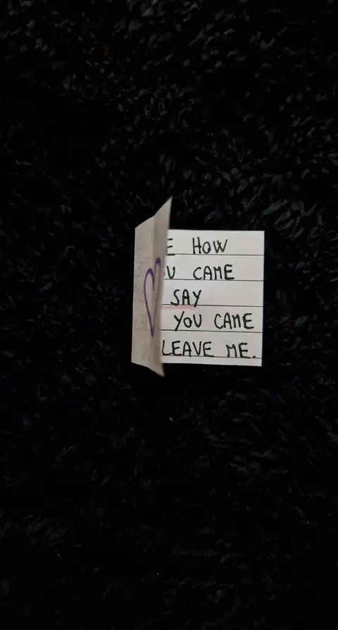 The image is a close-up shot of a piece of paper against a dark, textured background. The paper is folded, and handwritten text with a heart drawn on it is visible. The background appears to be a dark, possibly black, fuzzy surface.
