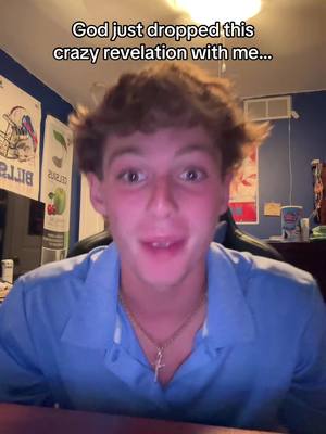 A young man with light brown hair is in a room, framed from the chest up. He has a neutral expression on his face, with his mouth slightly open. He's wearing a light blue polo shirt and a gold necklace with a cross pendant. In the background, there are posters and a lit ceiling fixture.