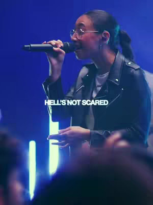 A woman with dark hair pulled back, wearing glasses and a black leather jacket, holding a microphone and singing. A blue background subtly fades in the background.
