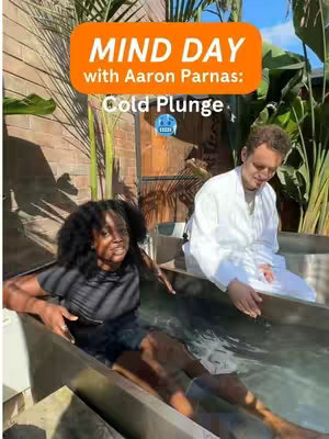 The cover image captures a outdoor scene with two people, a woman and a man, in a cold plunge tub. The woman, with dark curly hair, is seated in the water with a slight expression of surprise or focus. The man, wearing a white robe, is seated next to the tub in front of a building. The background shows palm trees and other foliage, suggesting a natural surrounding. An orange oval overlaid in the center contains the text as hook.