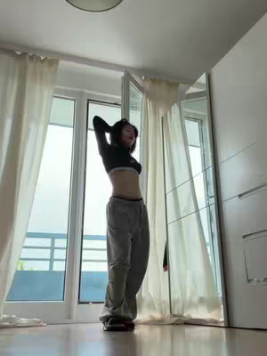 A young woman stands with her arms behind her head, wearing a black crop top and gray sweatpants. She is in a bright room with sheer curtains and a large window behind her, tilting her head back slightly, with natural side lighting and sunlight flooding the room. A mirror is visible to the right.