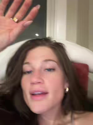 A selfie-style shot of a young woman lying down. Her head and shoulders fill the frame. She has brown hair, blue eyes, and is looking towards the camera. She's wearing hoop earrings and a ring. The background is out of focus, with a hint of a window and a red/brown surface, possibly a headrest.